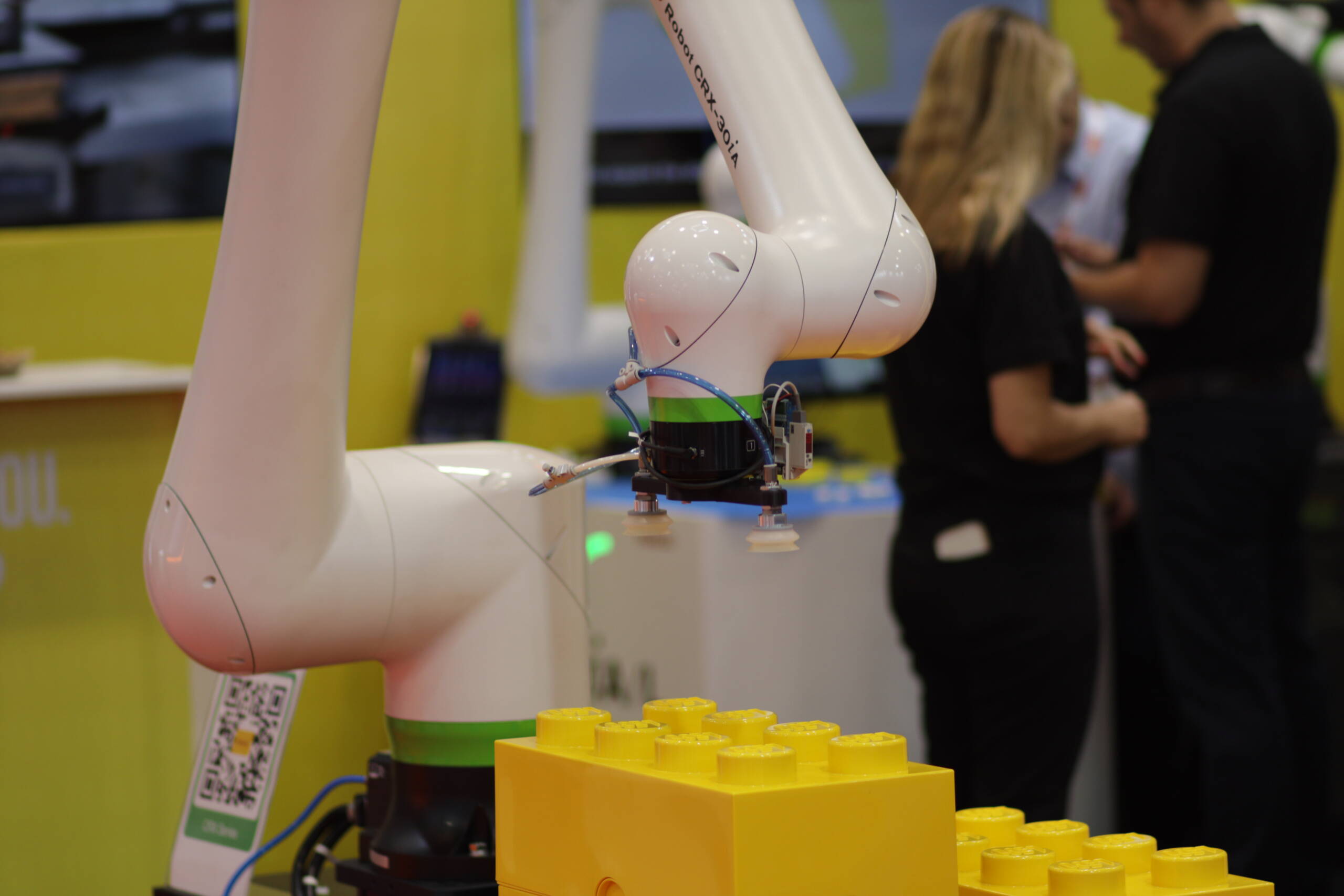 A collaborative robot arm with a vacuum-suction end effector picks up an oversized yellow block at an automation trade show demo